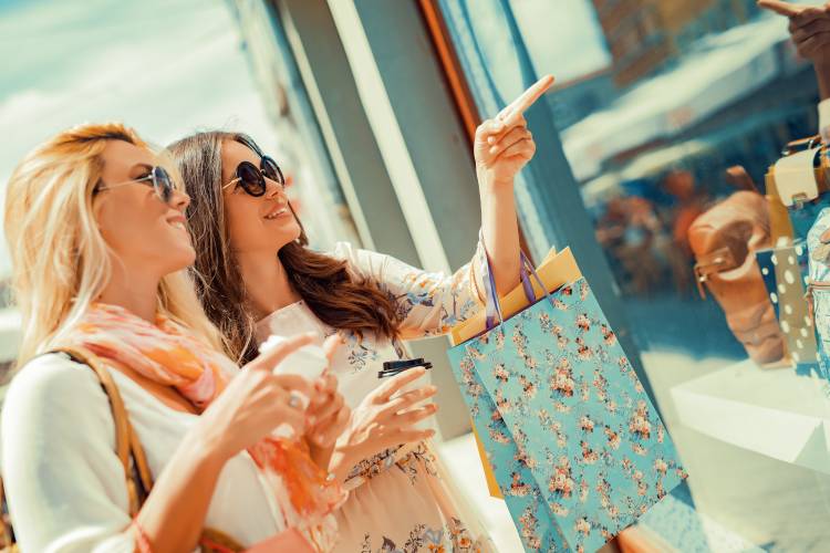 Two women holding shopping bags while looking in a shop window from outside on a sunny day