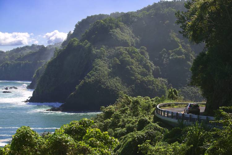 Wide view of a car making a sharp turn on the Road to Hana in Maui. The ocean and mountains are seen to the left.